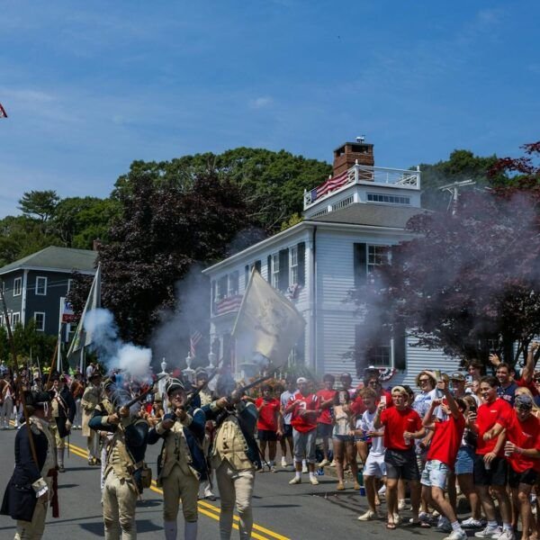 Crowded parade with reenactors in uniform firing rifles, surrounded by festive people waving flags on a sunny day.